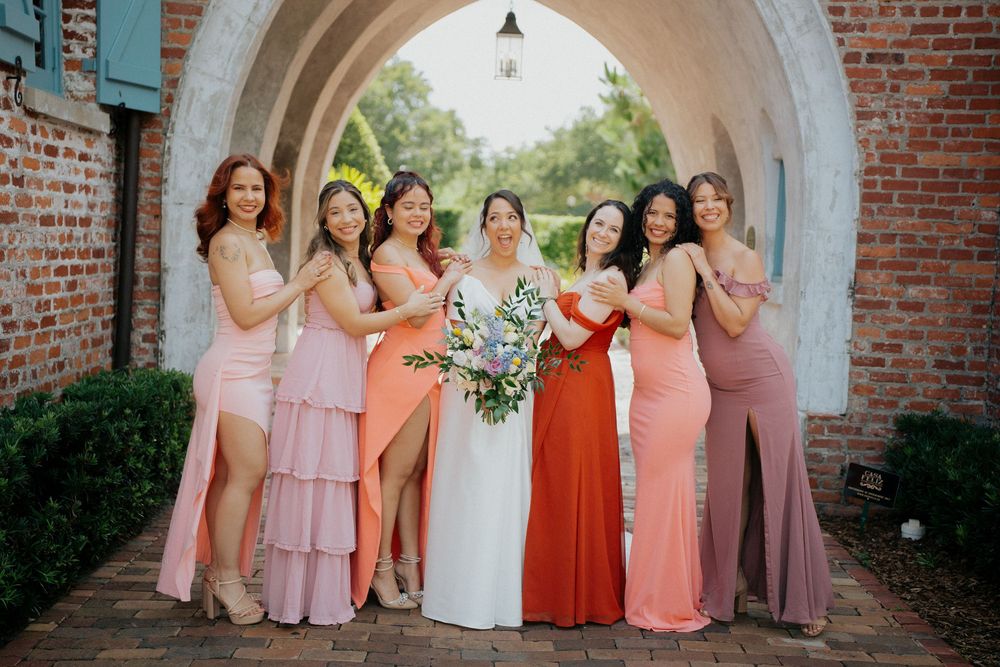 A bridal party poses under a brick archway wearing various shades of pink and coral dresses with a bride in white.