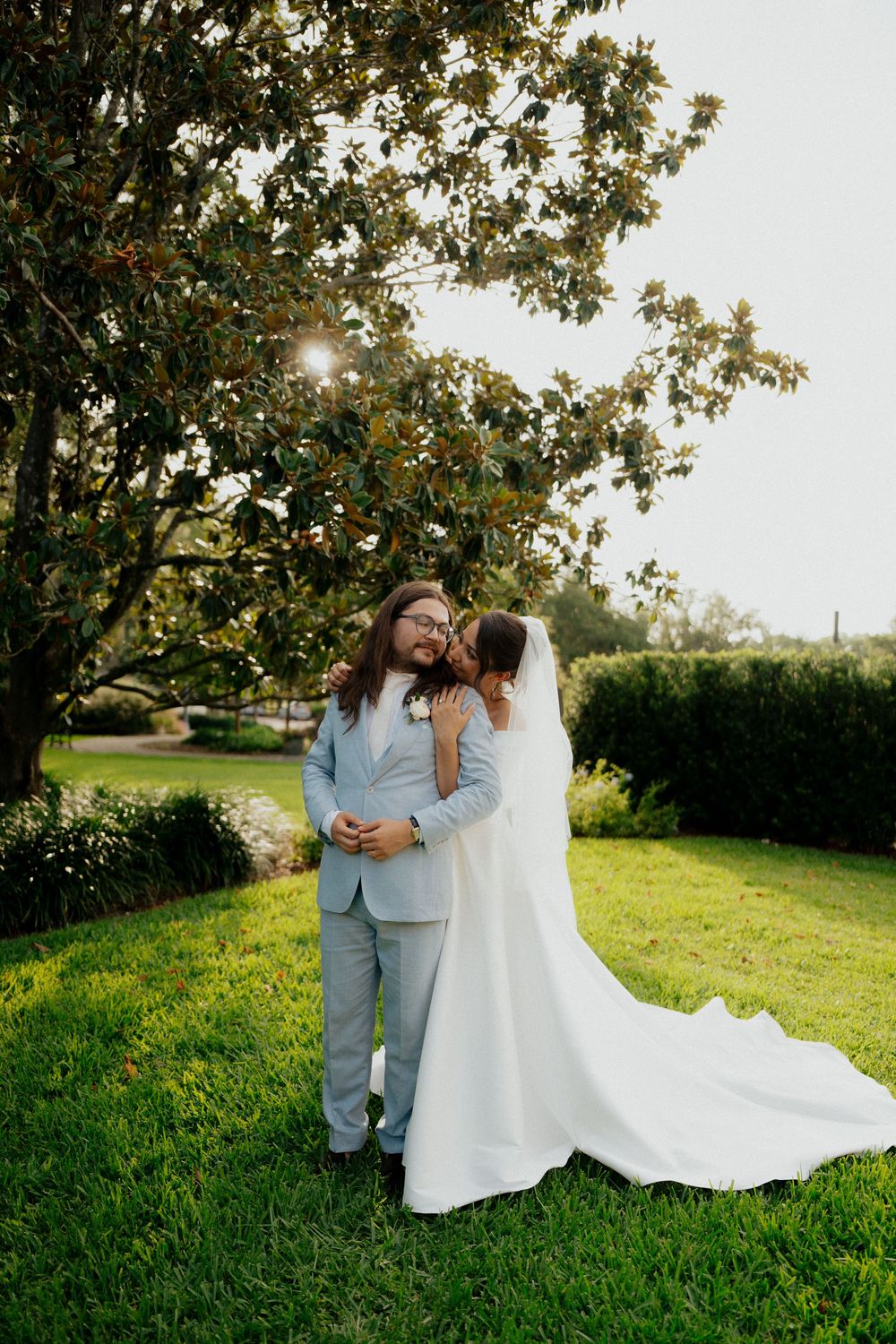 A romantic wedding photoshoot in a sunlit grassy field with trees creating a natural backdrop.