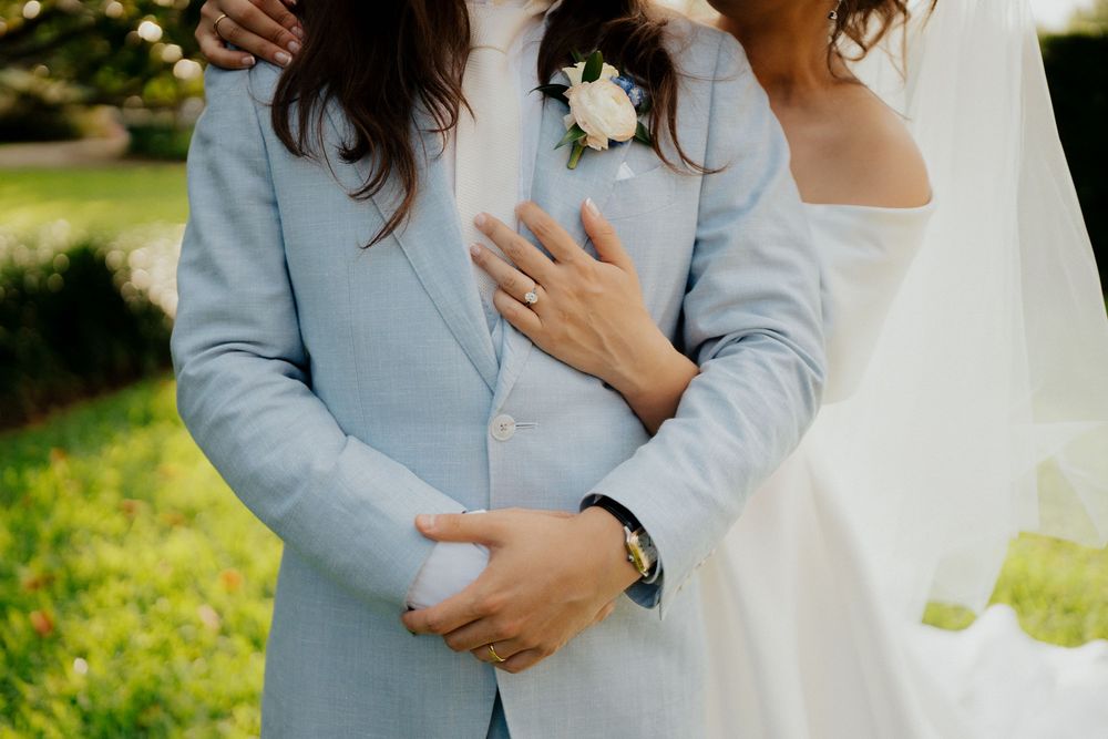 A couple in light blue and white wedding attire embrace outdoors in a romantic garden setting.