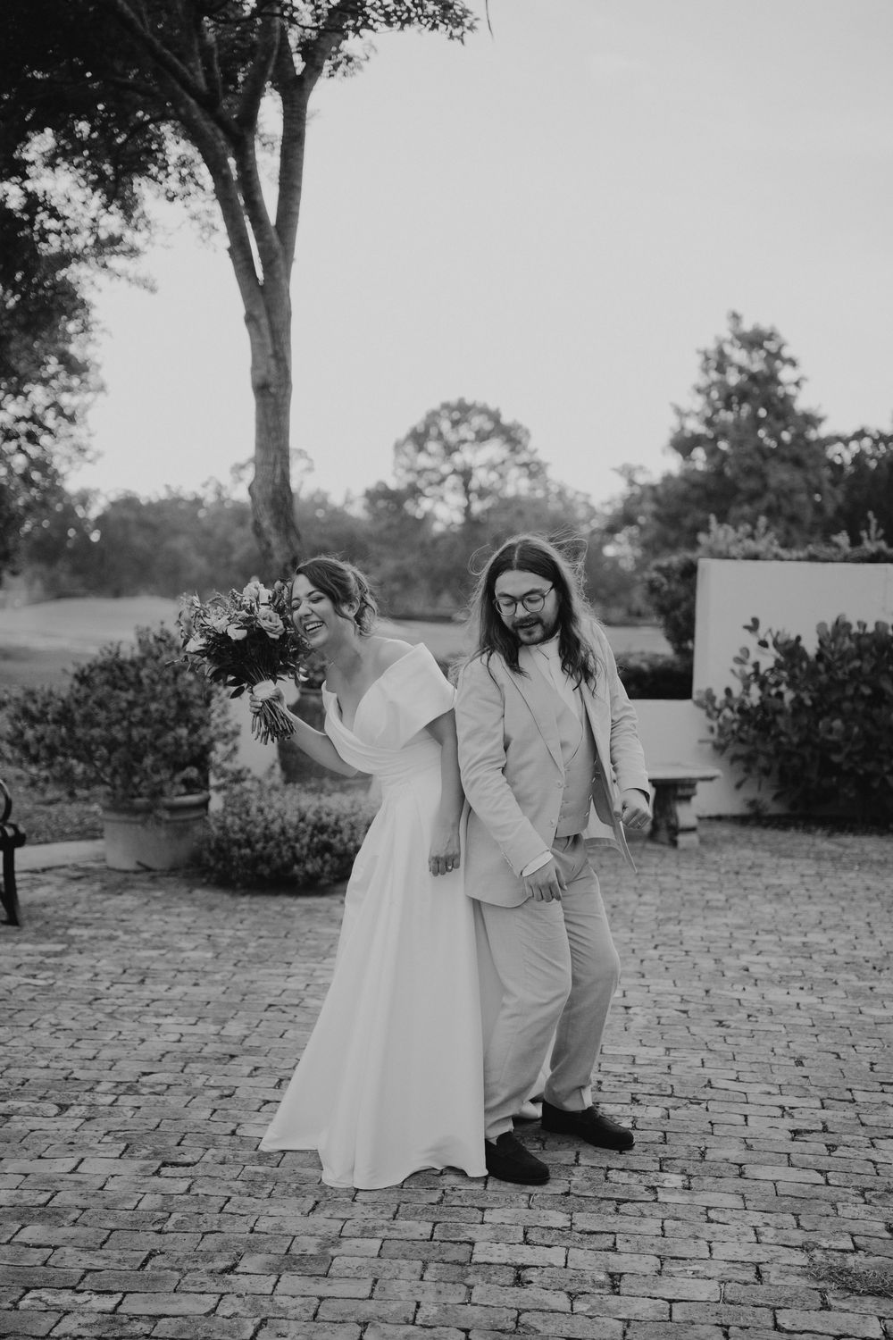 A black and white wedding photo series showing a couple posing on a brick patio under trees.