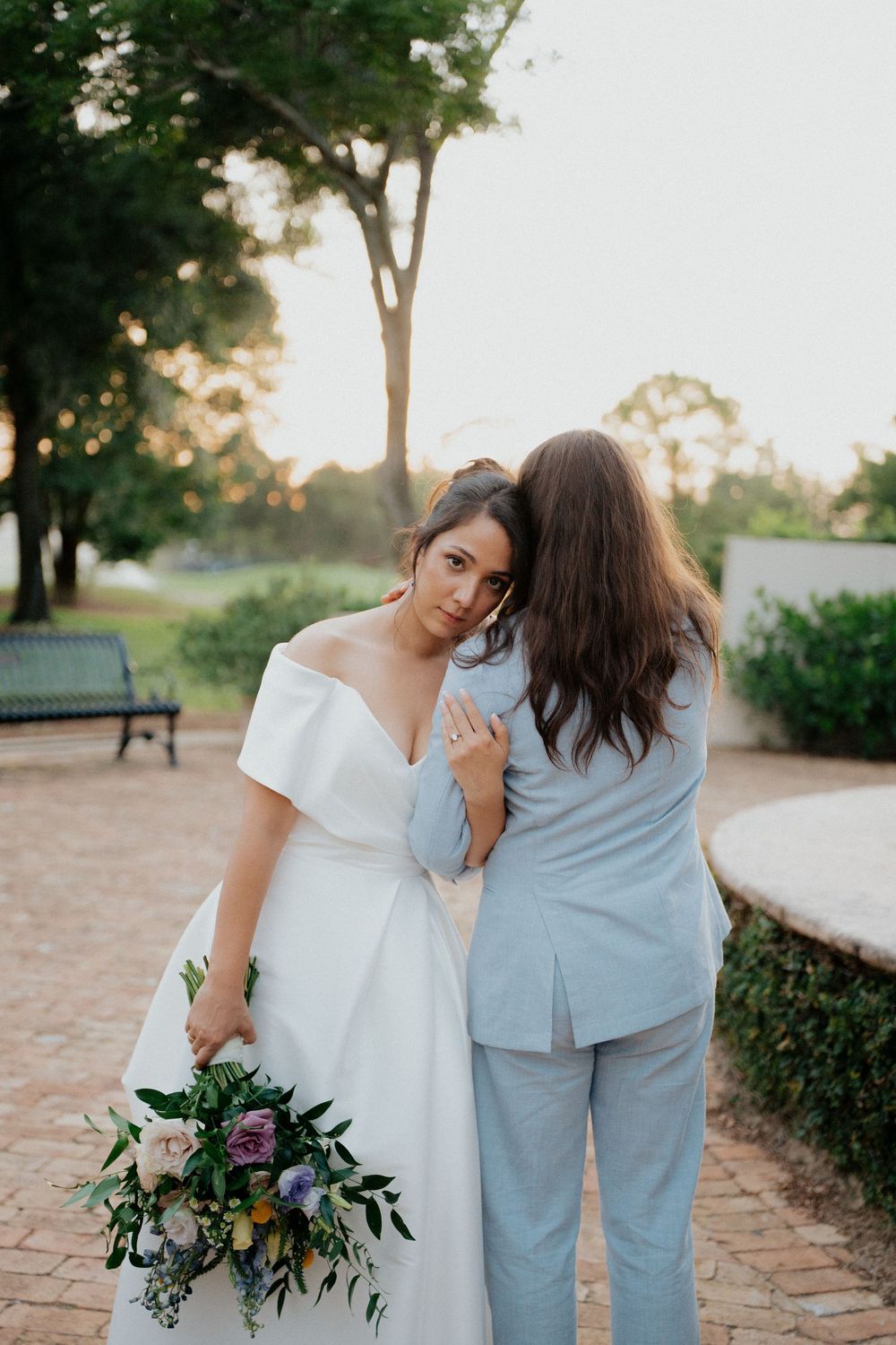Two people in coordinating white and blue outfits embrace while holding floral bouquets in a garden setting at sunset.