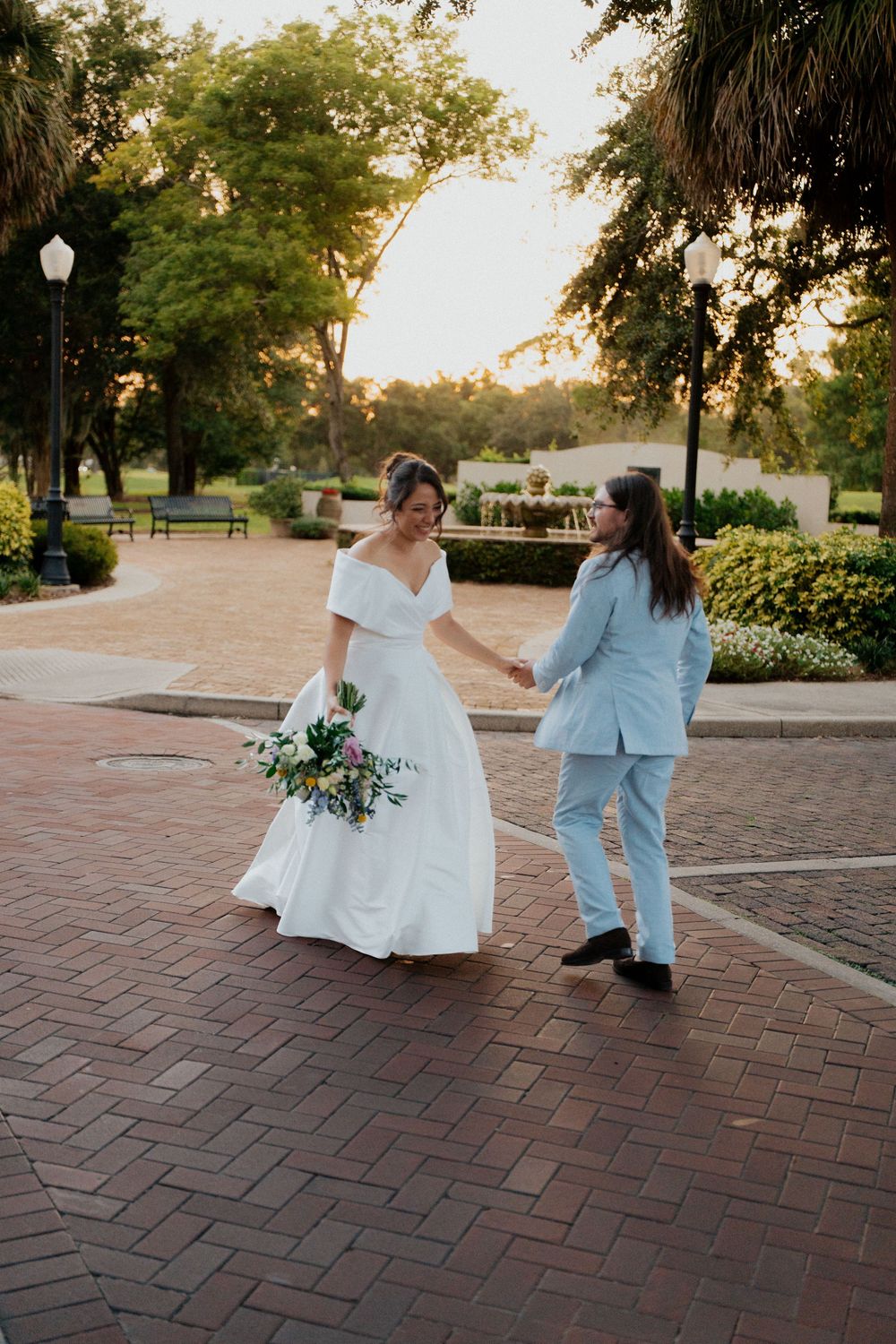 Two people in white and blue outfits dance playfully on a brick patio at sunset surrounded by palm trees and greenery.