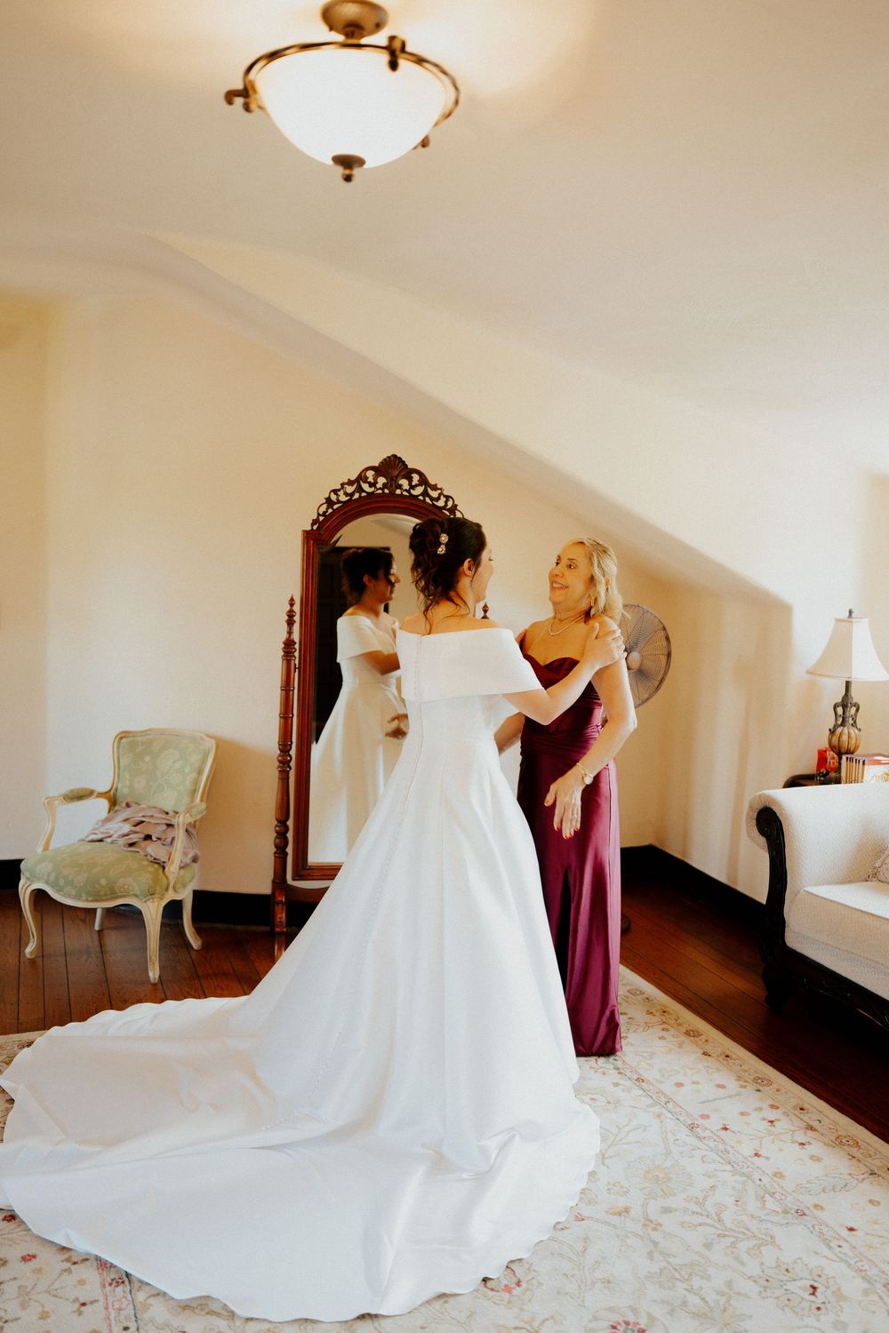 A bride in a flowing white wedding gown gets ready with help from attendants in a sunlit bridal suite.