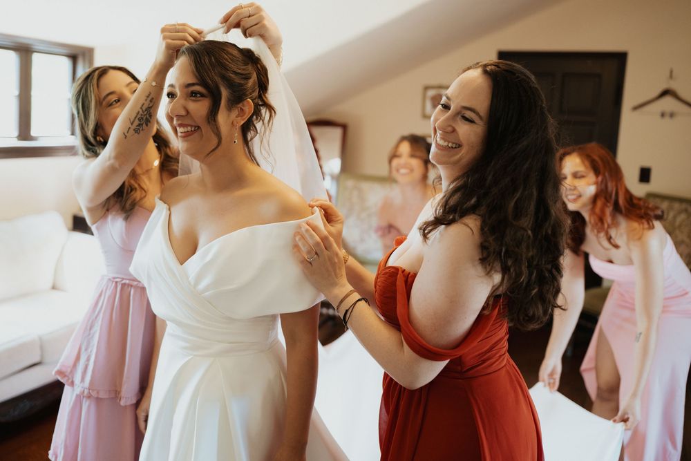 Wedding party getting ready in a bright room with bridesmaids in pink and red dresses helping with final preparations.