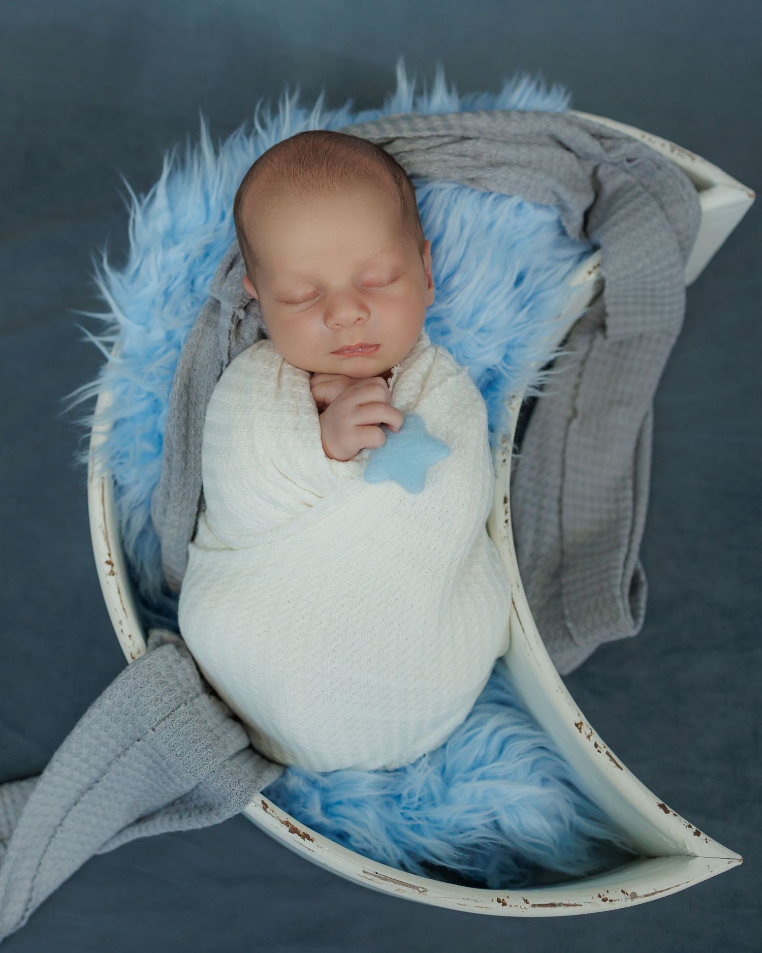 A sleeping newborn swaddled in white fabric rests in a curved bassinet with blue furry trim and gray padding.