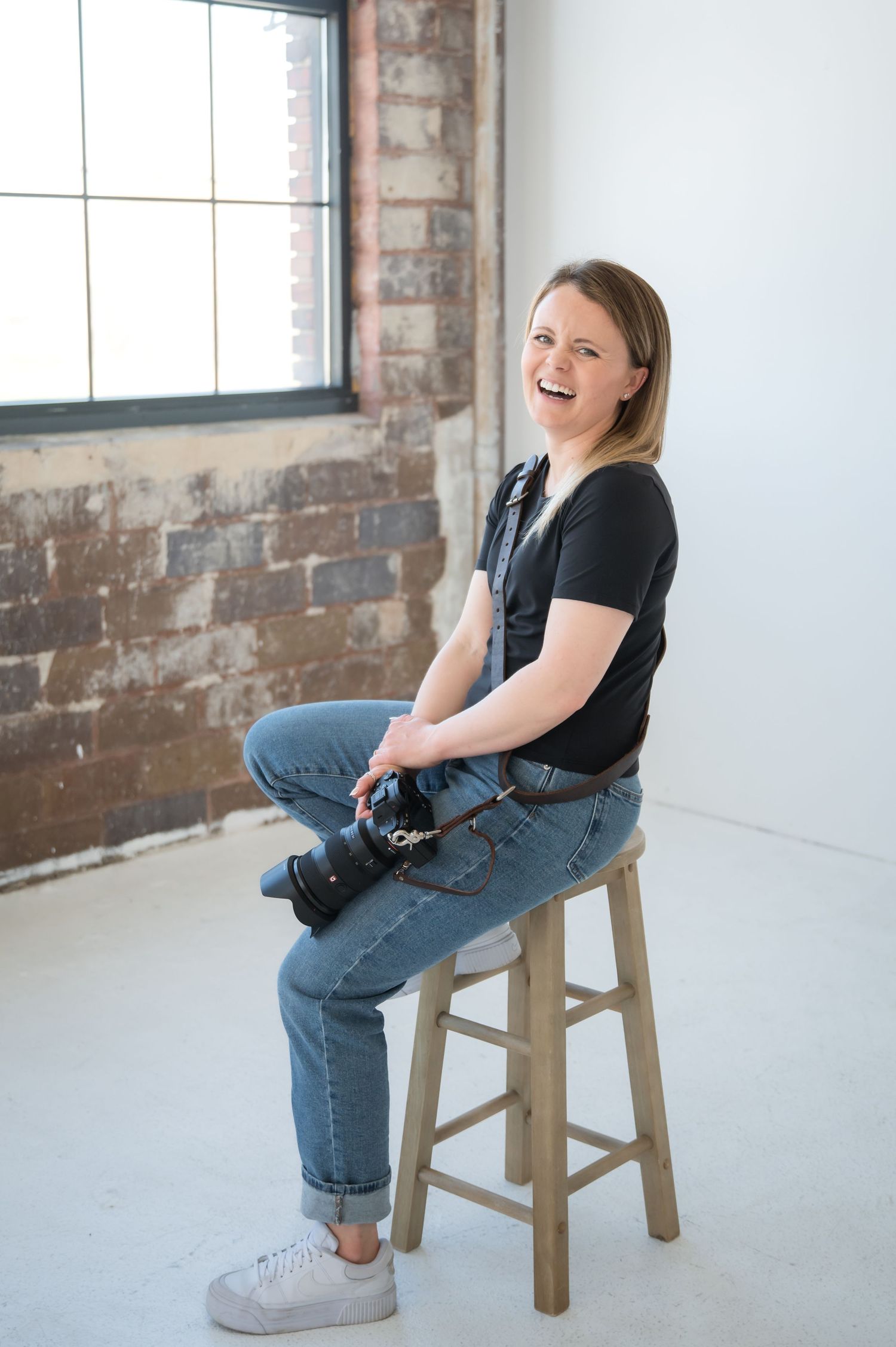 Person wearing black t-shirt and jeans sits casually on wooden stool in industrial loft space with exposed brick walls.