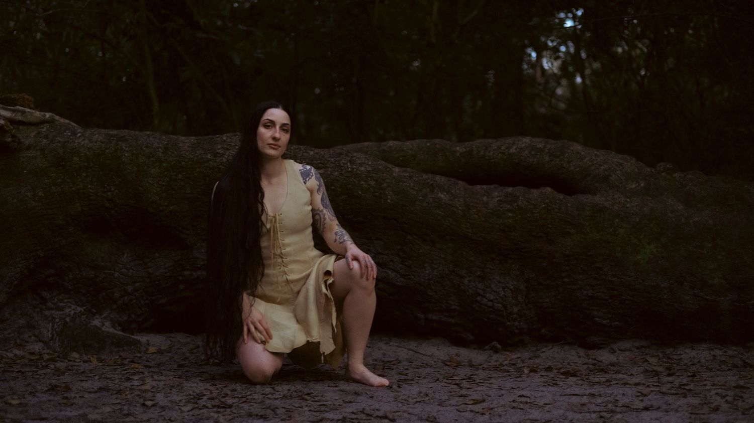 A person in a white dress sits on a rocky beach near the water during sunset.