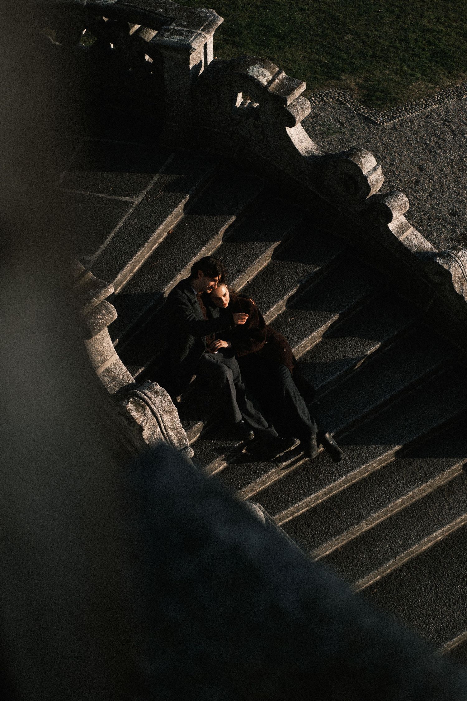 A couple in dark clothing sits together on stone steps during golden hour sunlight.