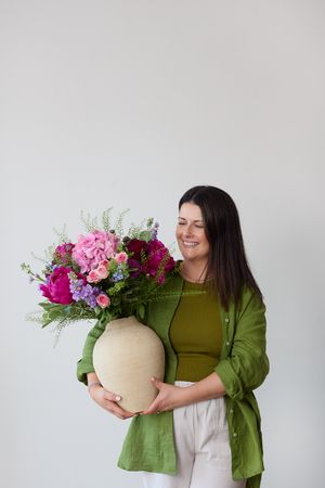 A woman in a green outfit holds a beige vase filled with vibrant pink and purple flowers against a white background.