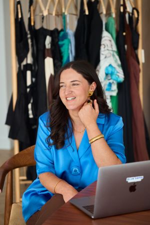 Fashion Business owner in blue satin shirt working on laptop with clothing rack in background. Personal Branding Photography.