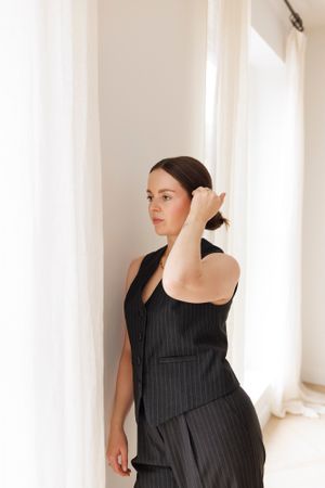 Elegant pose in black pinstripe outfit by sunlit window with flowing curtains. Personal Branding Photography.