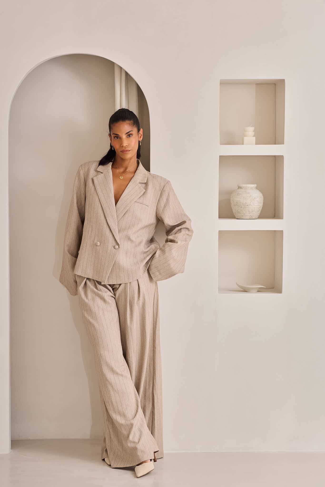 Minimalist beige pantsuit styled with white vases in wall niches against neutral background.