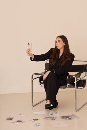 Model in black outfit poses artistically on modern chrome chair against beige backdrop with scattered photos.