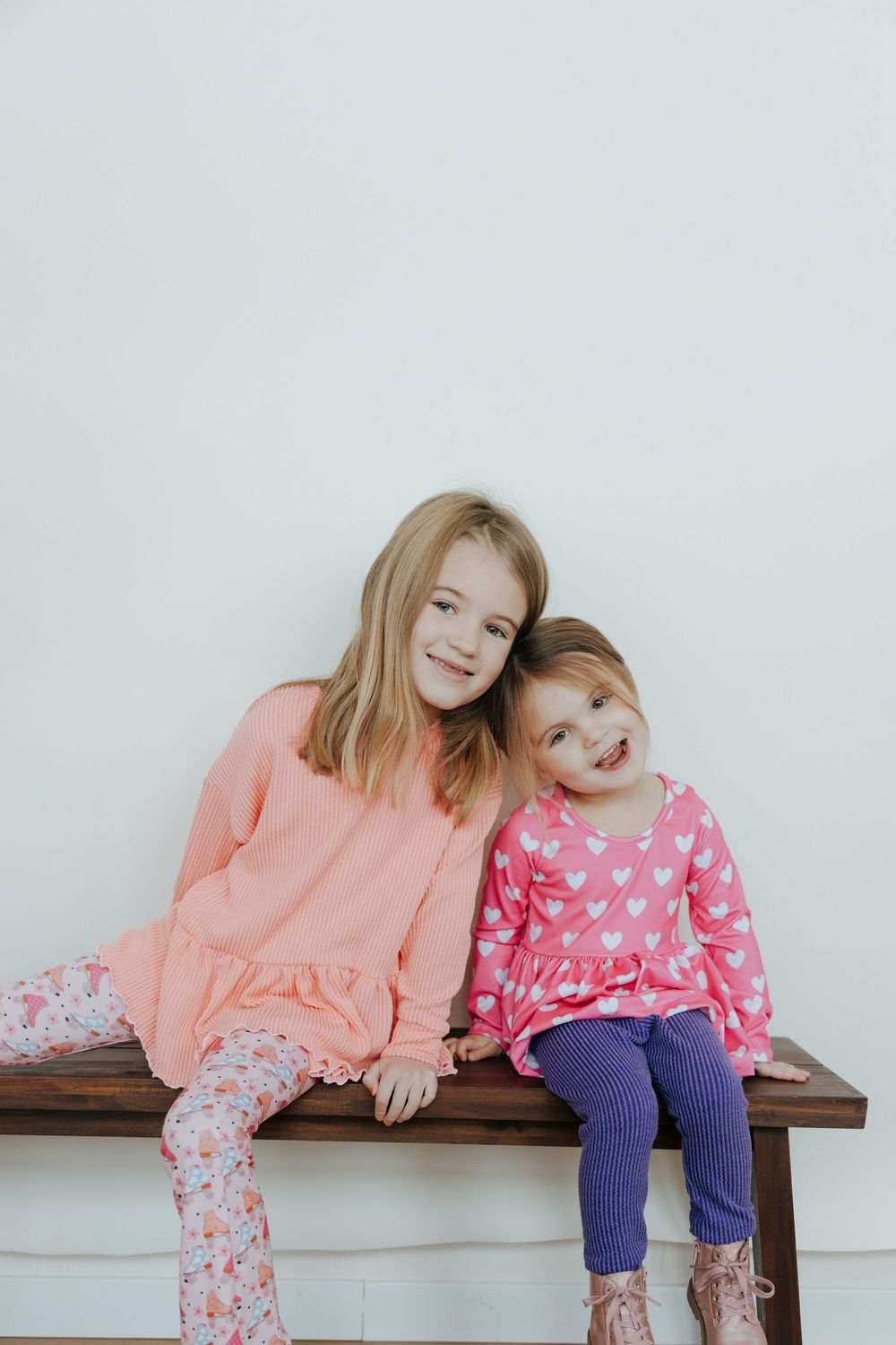 Two children in matching pink and coral outfits sit together on a wooden bench against a white wall.