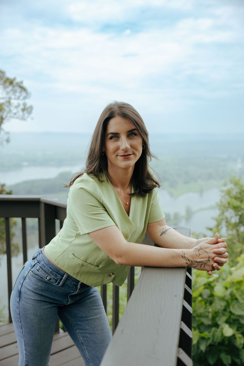 Person in light green shirt and jeans leans on white railing of outdoor deck with scenic mountain view in background.