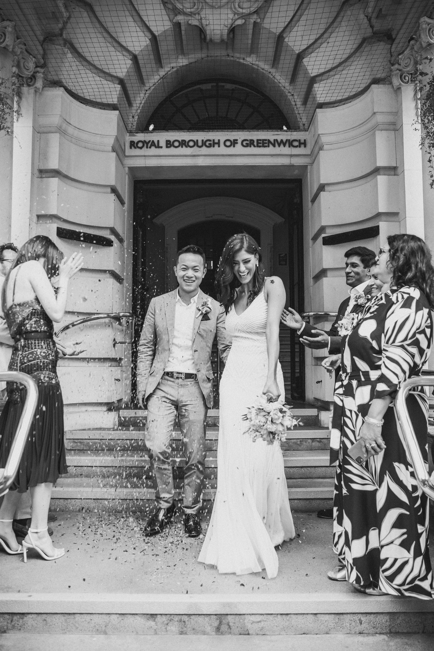 Newlywed couple exits Royal Borough of Greenwich registry office as guests celebrate by throwing confetti in black and white wedding photo.