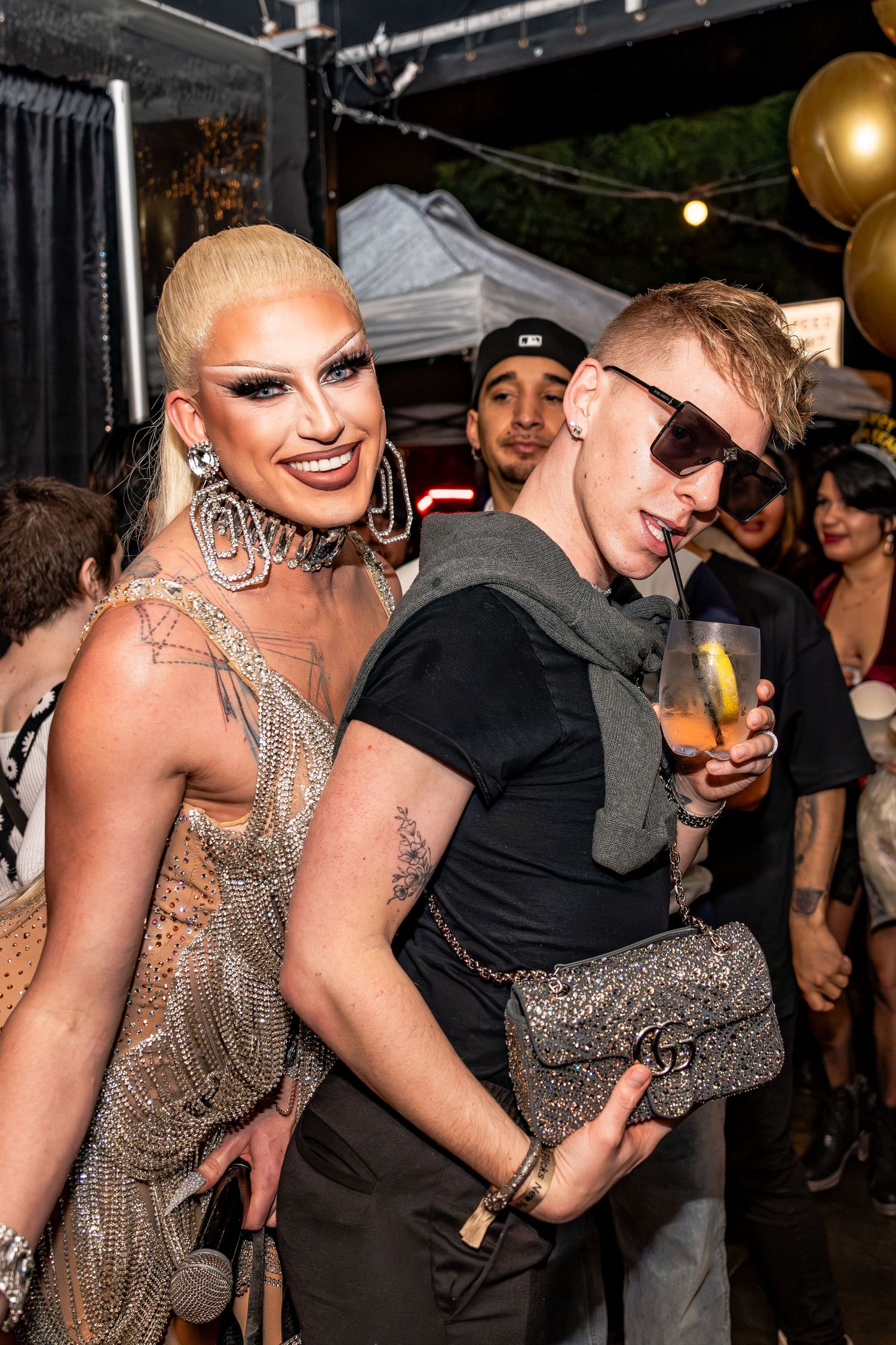 New Years Eve at Flicks: Drag queen Joey Jay in a bedazzled fringe dress stands with a blonde man sipping his drink looking at the camera.
