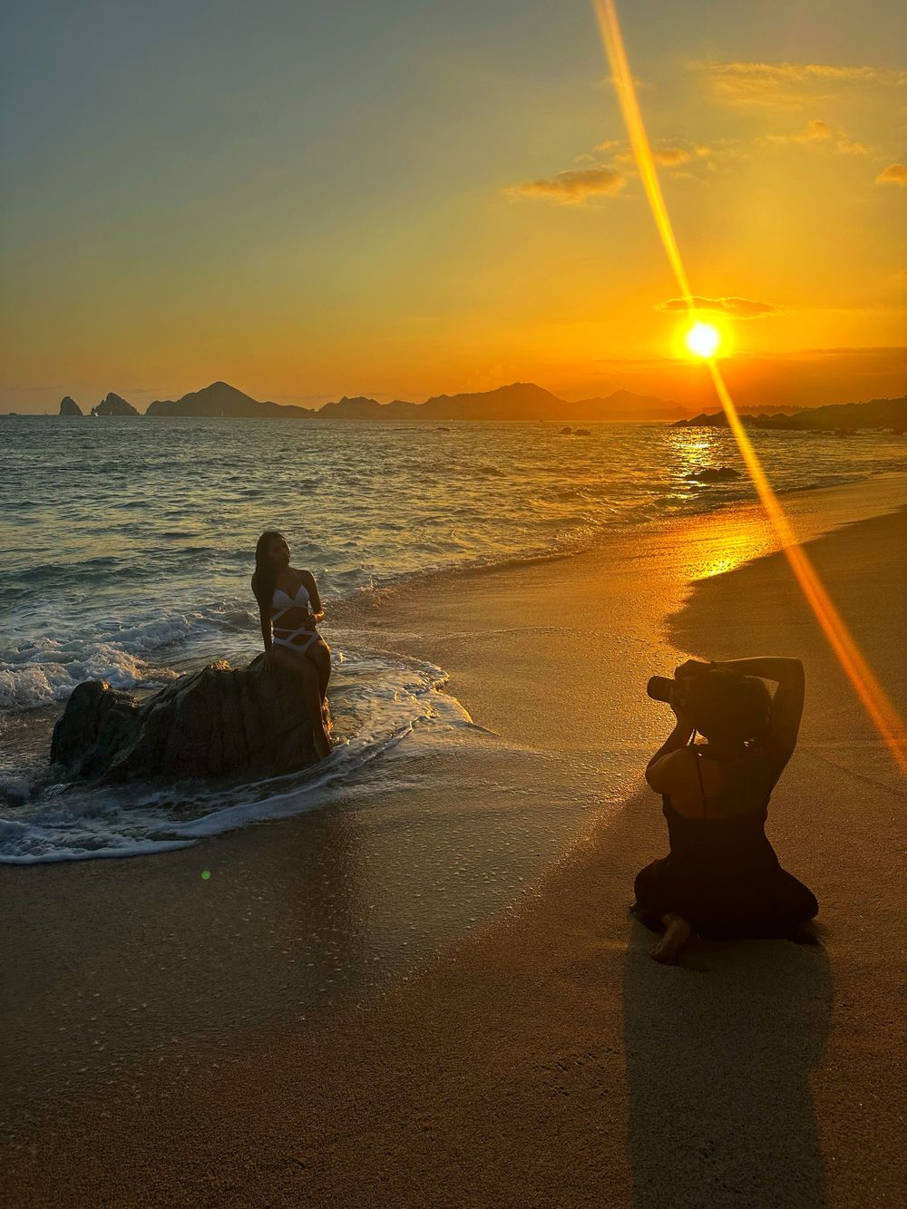 Silhouette of photographers capturing sunset on a beach with rocks and ocean waves.