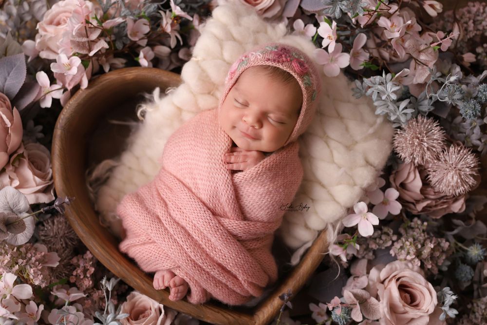 Sleeping newborn wrapped in pink knit blanket surrounded by delicate pink flowers and foliage.