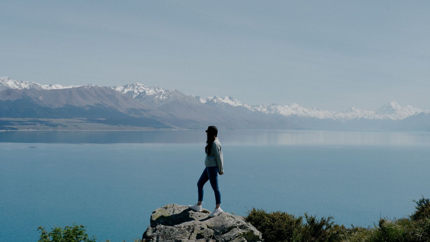 Silhouette standing on rocky outcrop overlooking vast blue lake and mountain range.