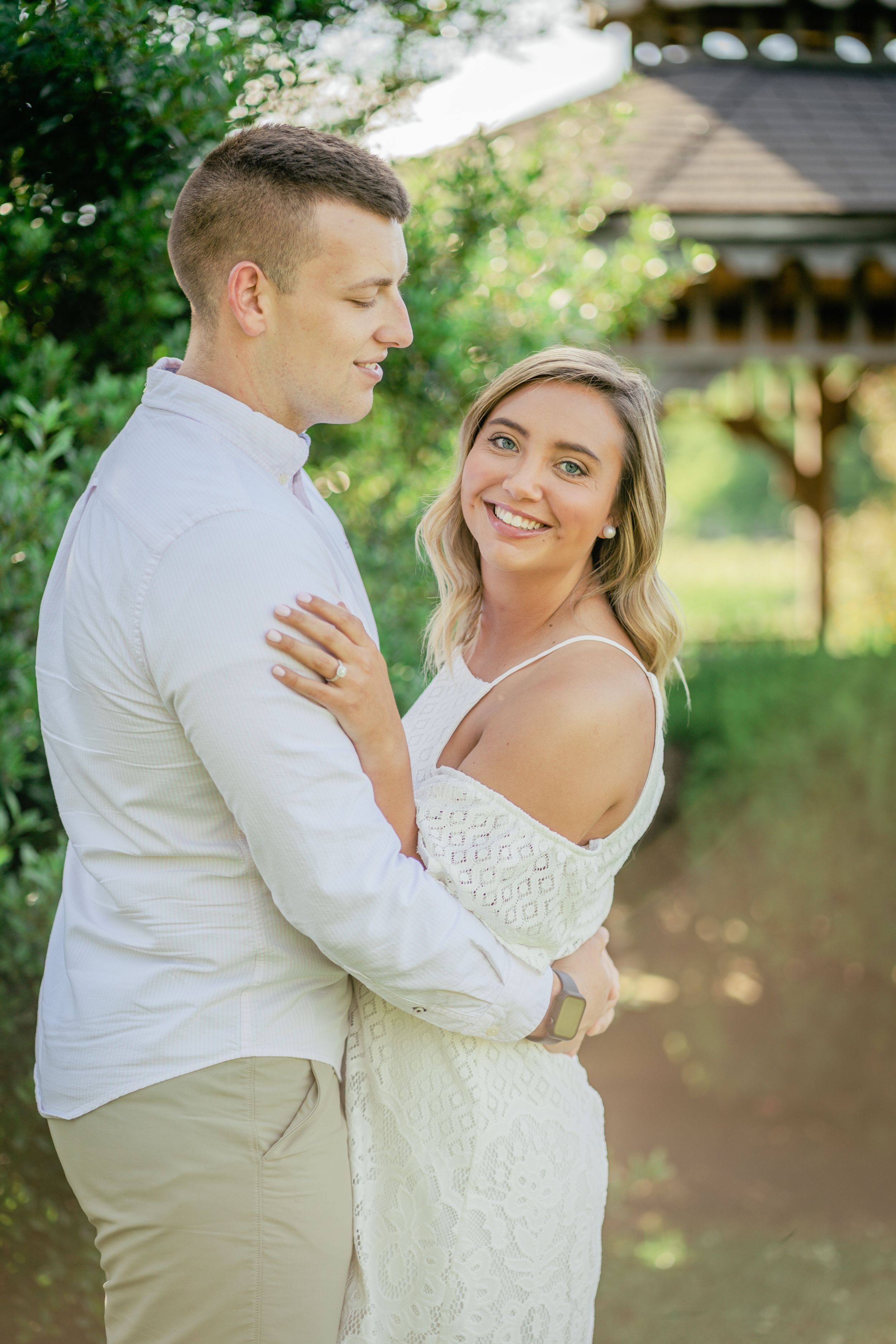Couple in white outfits embrace during engagement photoshoot in garden setting.