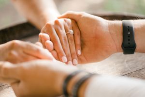 Close up of hands touching on a wooden surface, with one hand wearing an engagement ring and a black fitness tracker.