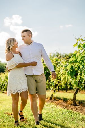 Couple walking together through sunny vineyard in casual white summer outfits.