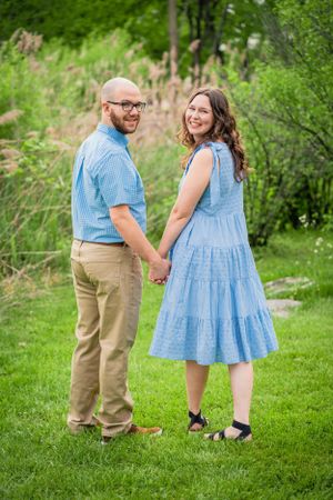 A couple stands holding hands on green grass with wildflowers and trees in the background.