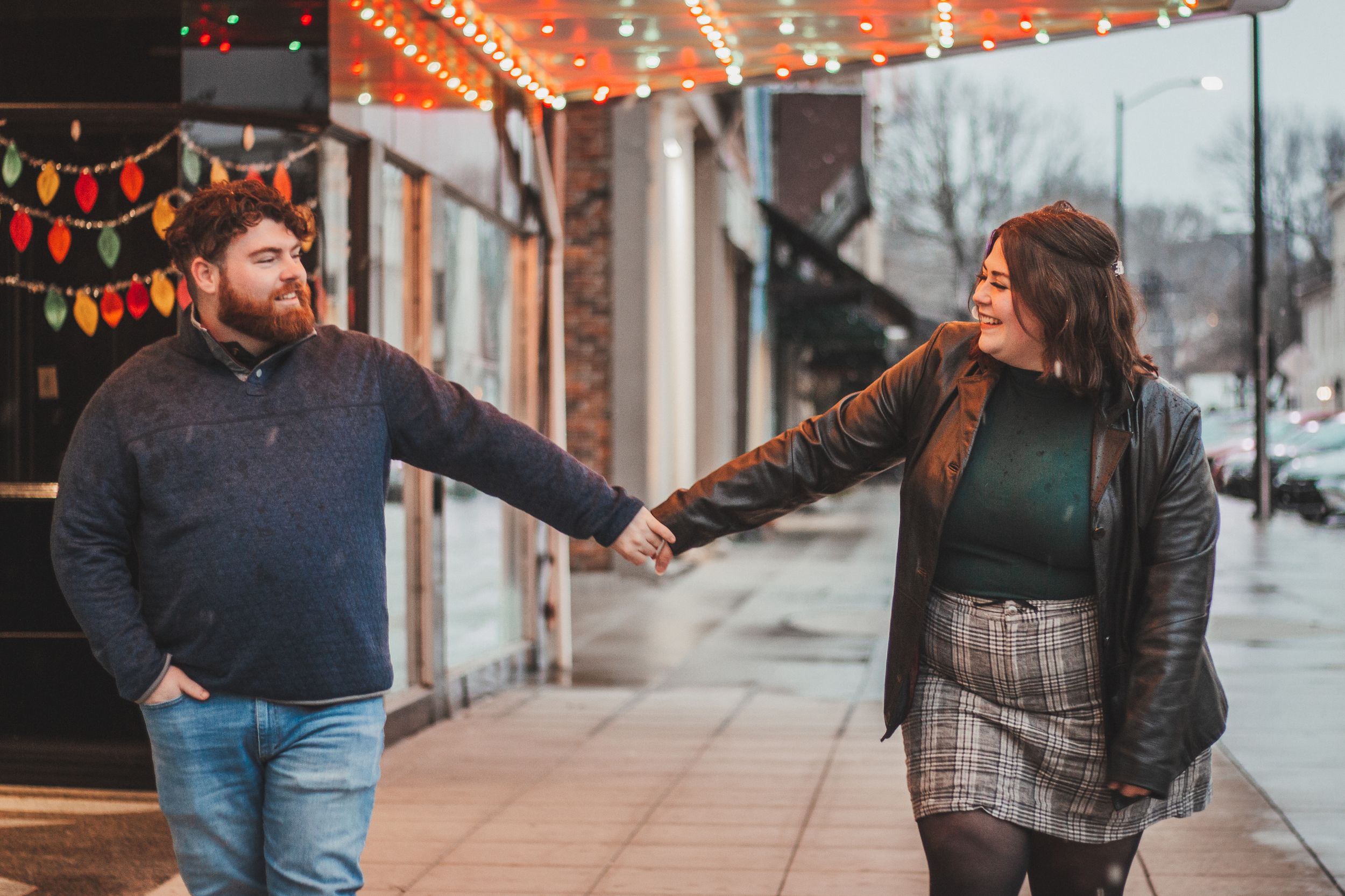 A couple playfully holds hands under festive string lights and colorful decorations.