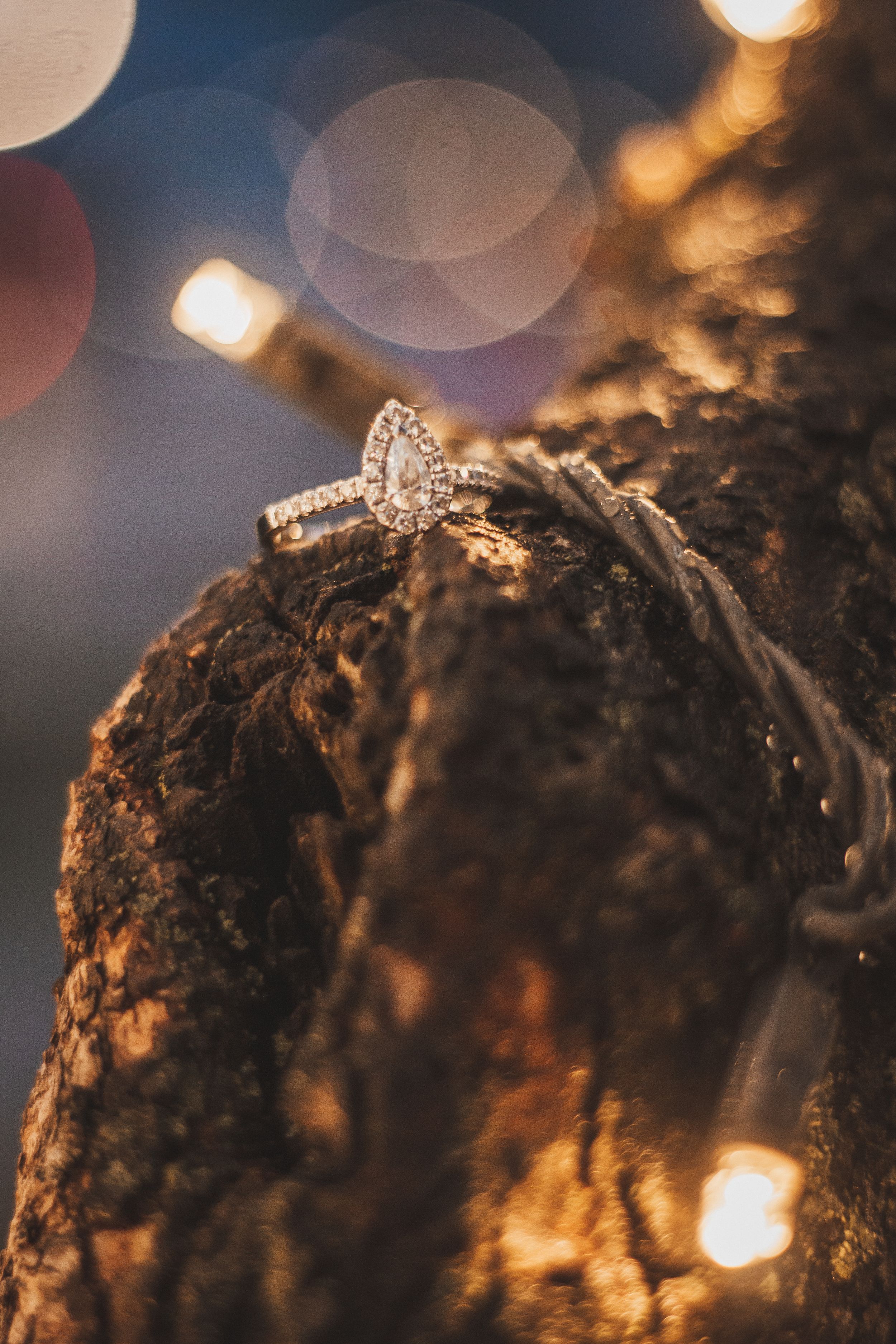 A pear-shaped diamond engagement ring sparkles against a textured surface with bokeh lighting effects.