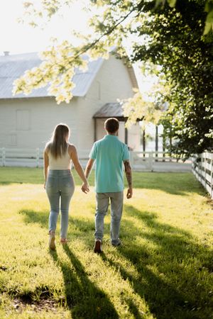Couple holding hands while walking away towards white farmhouse at sunset.