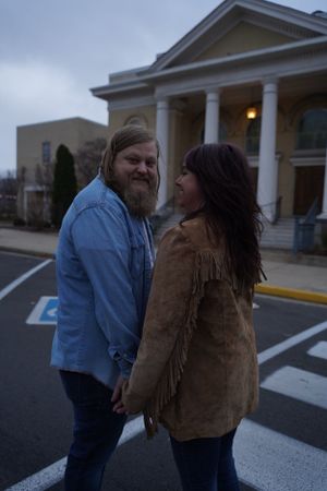 A couple shares a tender moment in a parking lot outside a building with columns at twilight.
