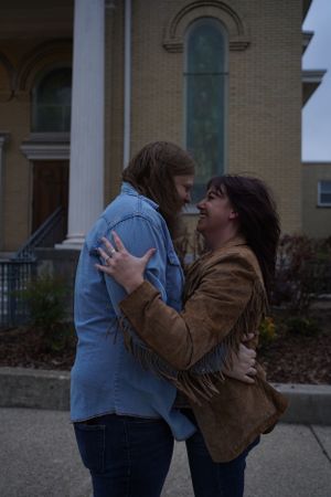 A couple embraces outside a historic brick building with arched windows at dusk.