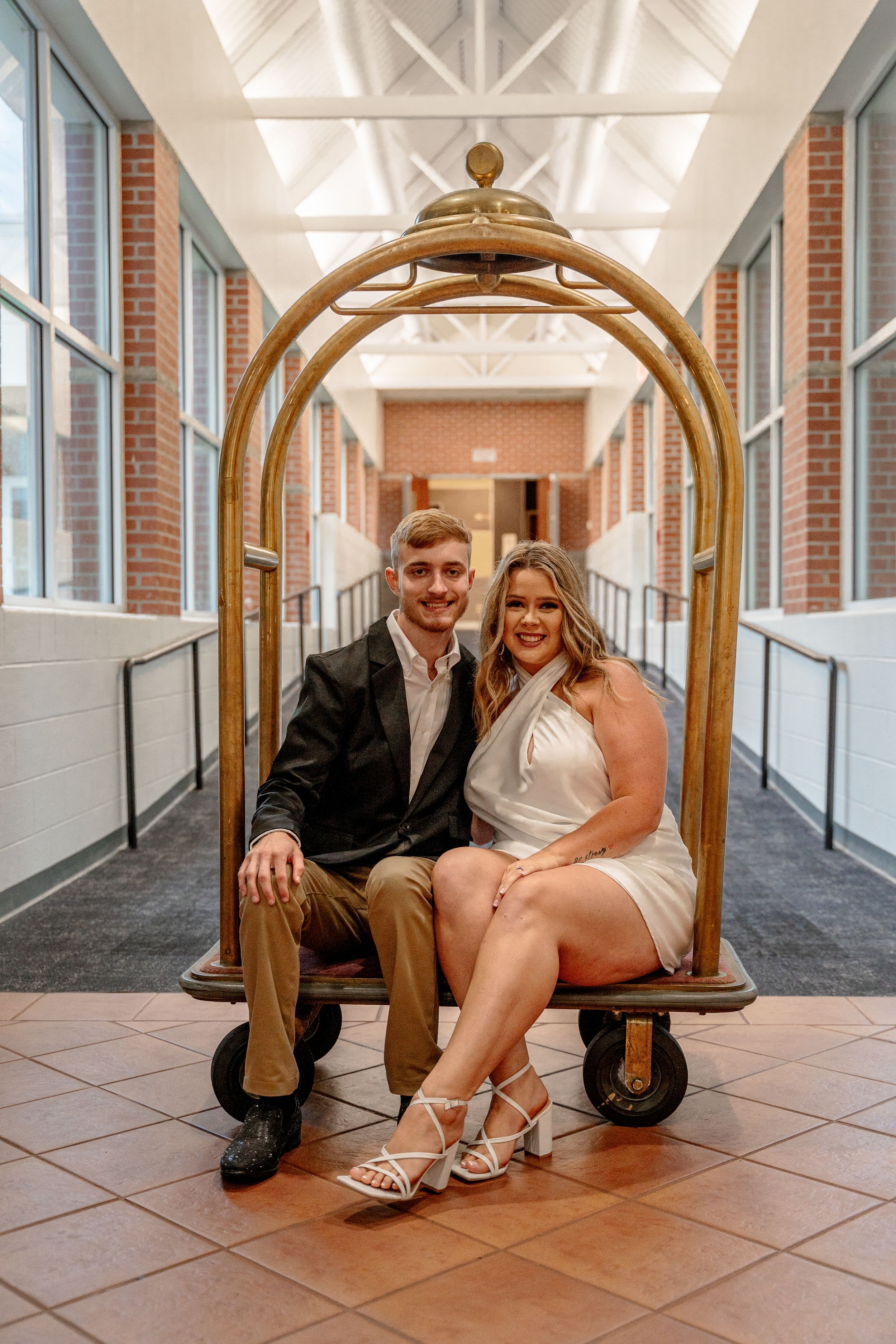 Couple posing together on antique luggage cart in elegant hallway.
