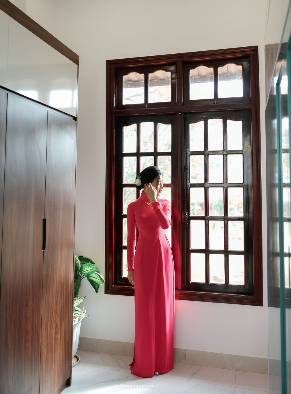 Elegant figure in red traditional dress standing by window with wooden frame interior.