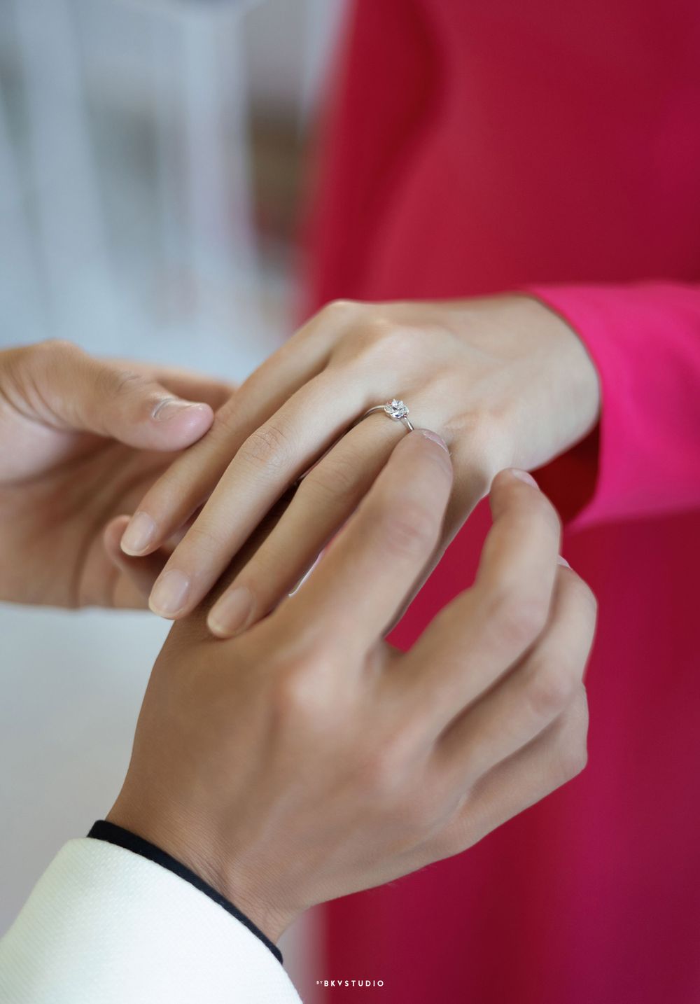 Close-up of hands exchanging an engagement ring with pink and white clothing visible.