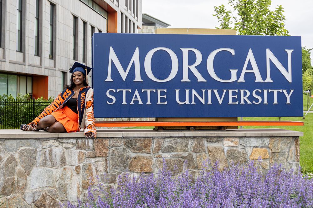 Graduate in cap and gown posing beside Morgan State University sign, wearing orange and black regalia.