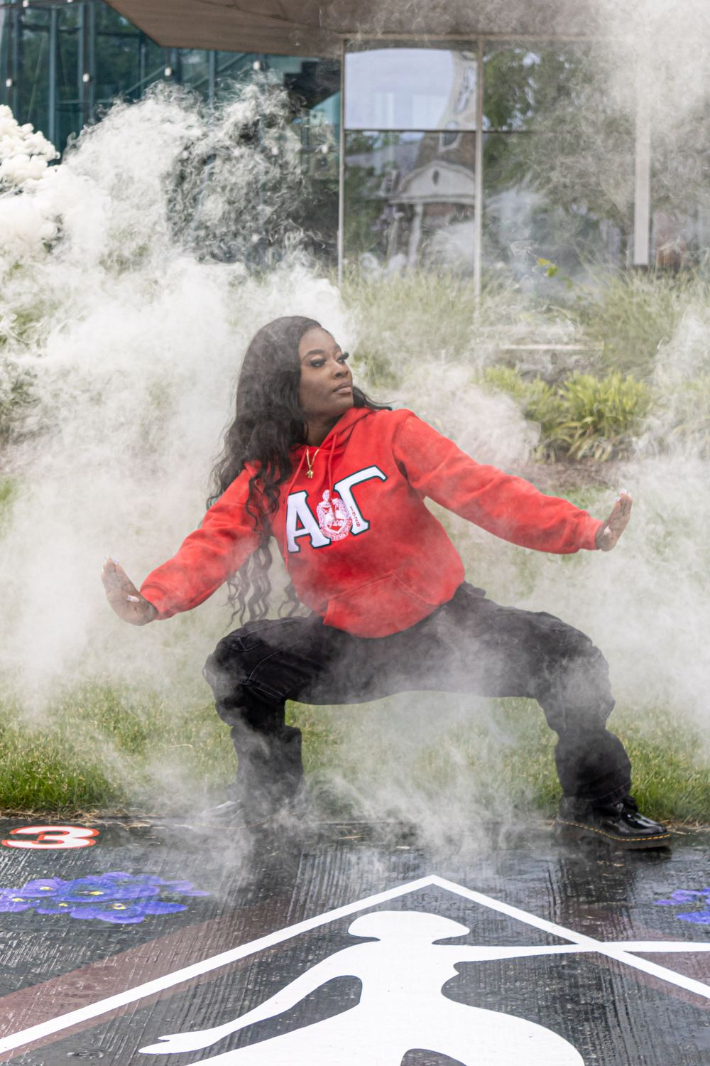 Woman in red Alpha Gamma hoodie strikes dynamic pose surrounded by white powder smoke outdoors.