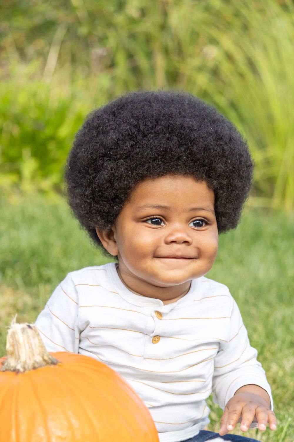 A toddler with an afro hairstyle sits next to a pumpkin outdoors with a bright smile.