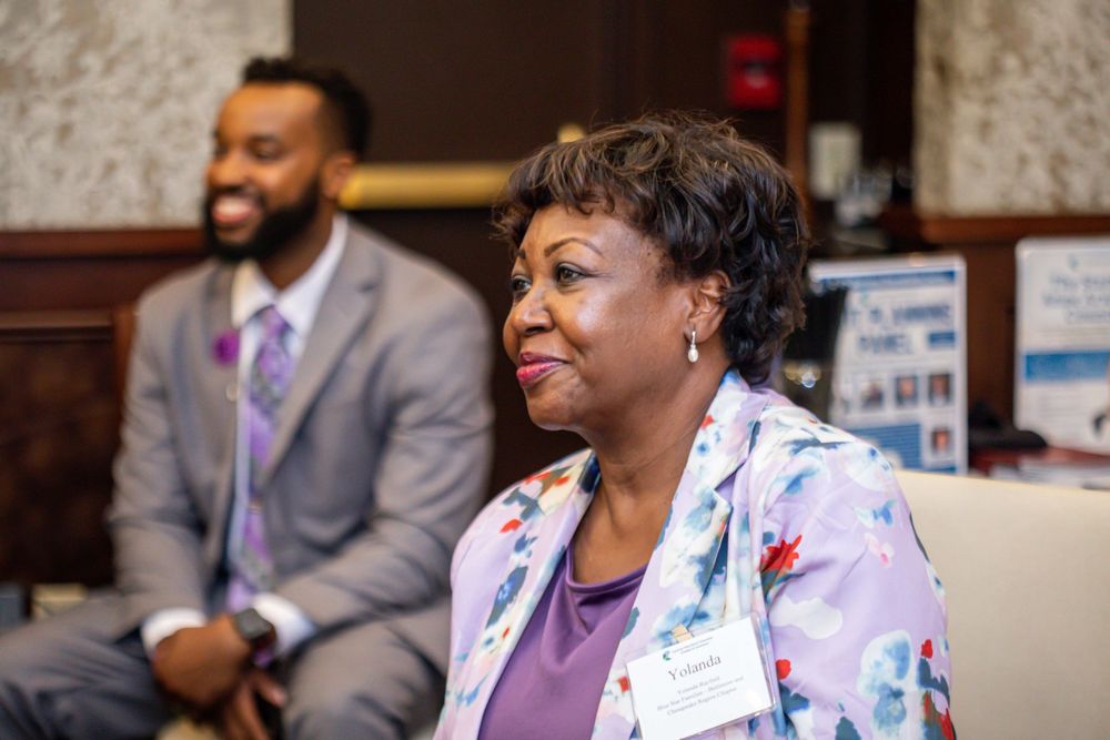 Two professionals in business attire smile at an indoor conference event, woman in floral blazer foreground.