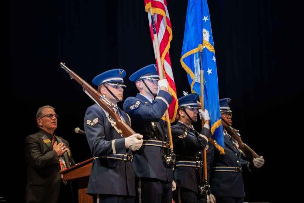 Military honor guard in blue uniforms presenting flags including US and Air Force flags at a formal ceremony.