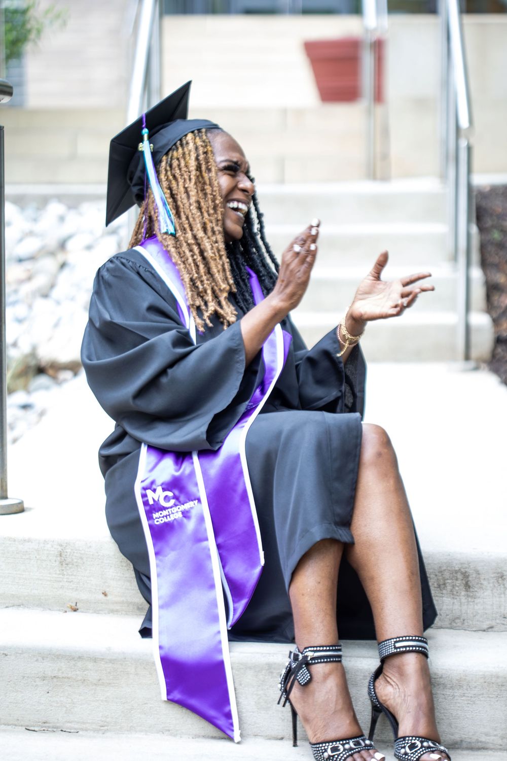 Joyful graduate in black cap and gown with purple sash sits on steps, clapping and smiling at graduation ceremony.