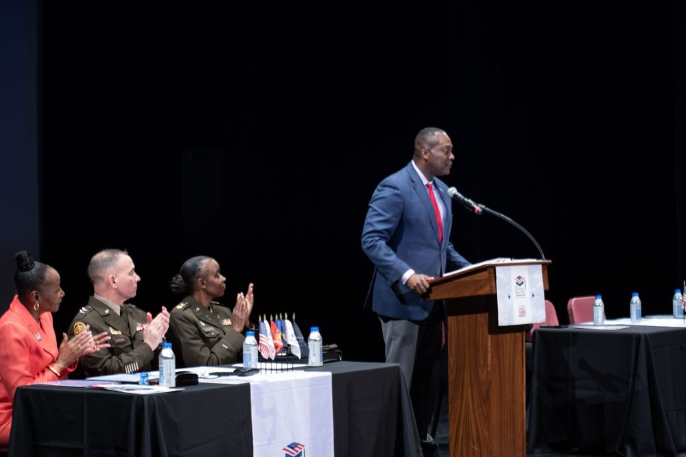 Man in blue suit speaks at podium during formal military panel event with officers seated at decorated table.