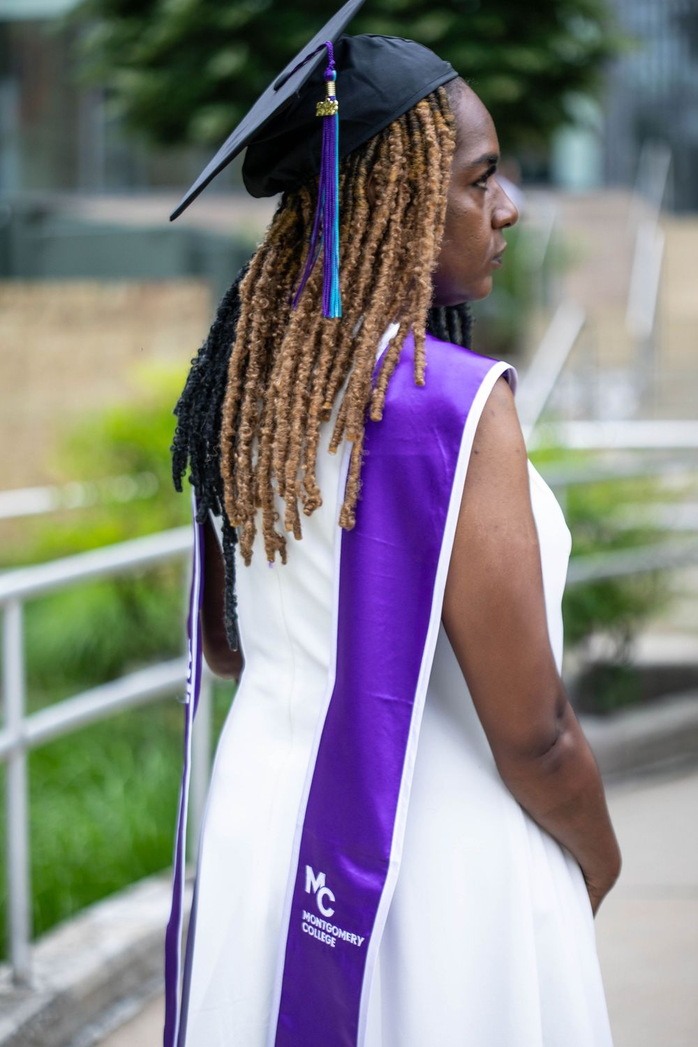 Graduate in white dress and purple Montgomery College sash poses outdoors wearing graduation cap with locs.