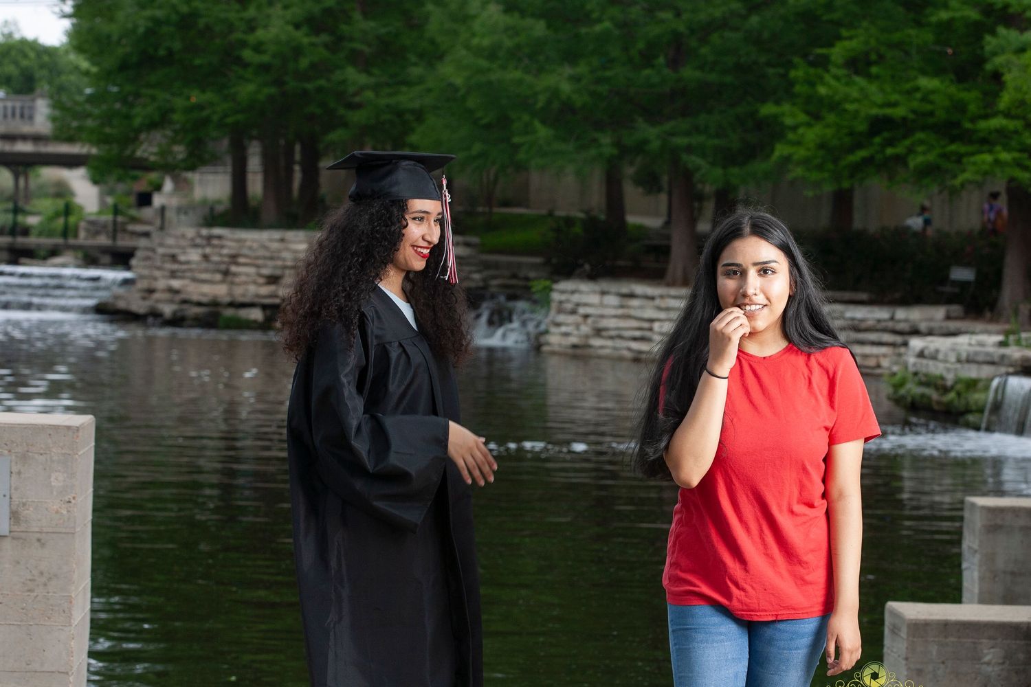 Two people posing by a stone wall near a pond, one wearing graduation regalia and one in casual attire.