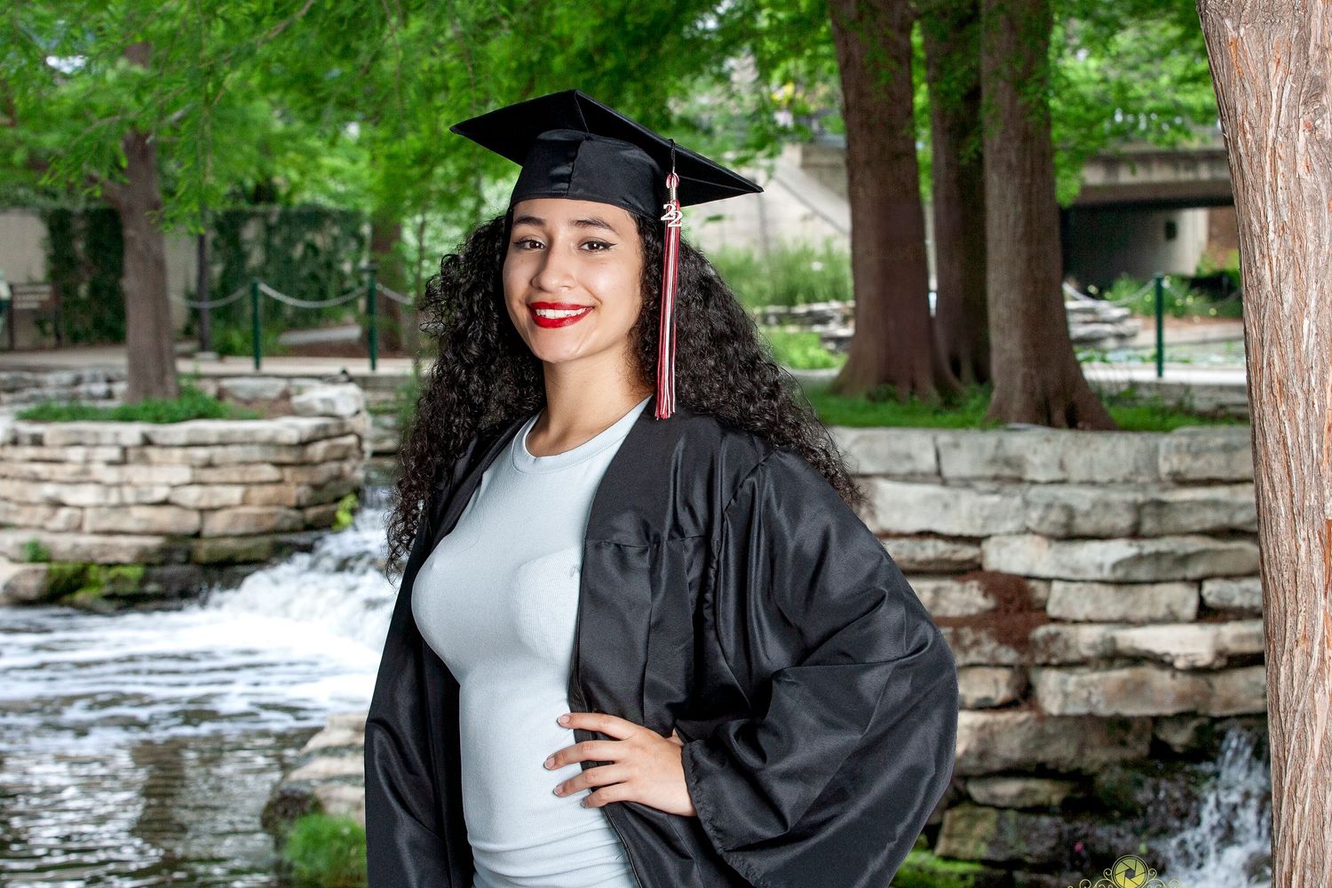 Graduate in black cap and gown poses confidently by stone wall with water feature in background.