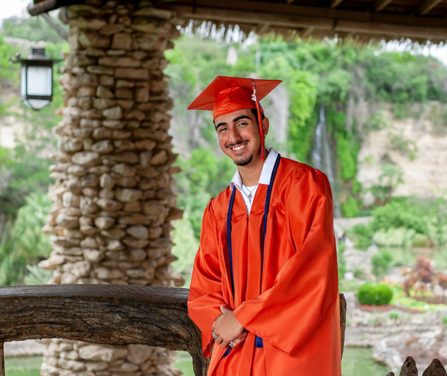 A graduate in red academic regalia stands under a stone pergola.