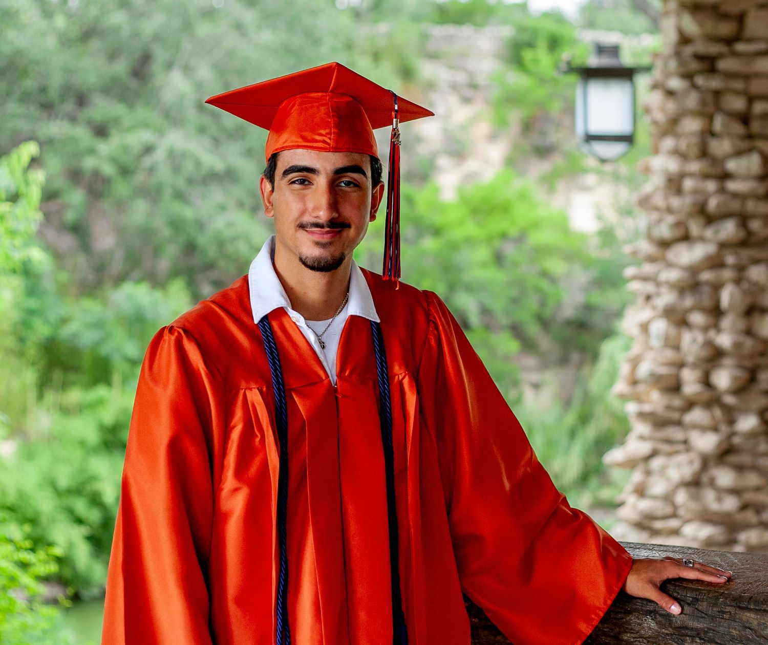 A graduate in bright red cap and gown poses under a stone archway.