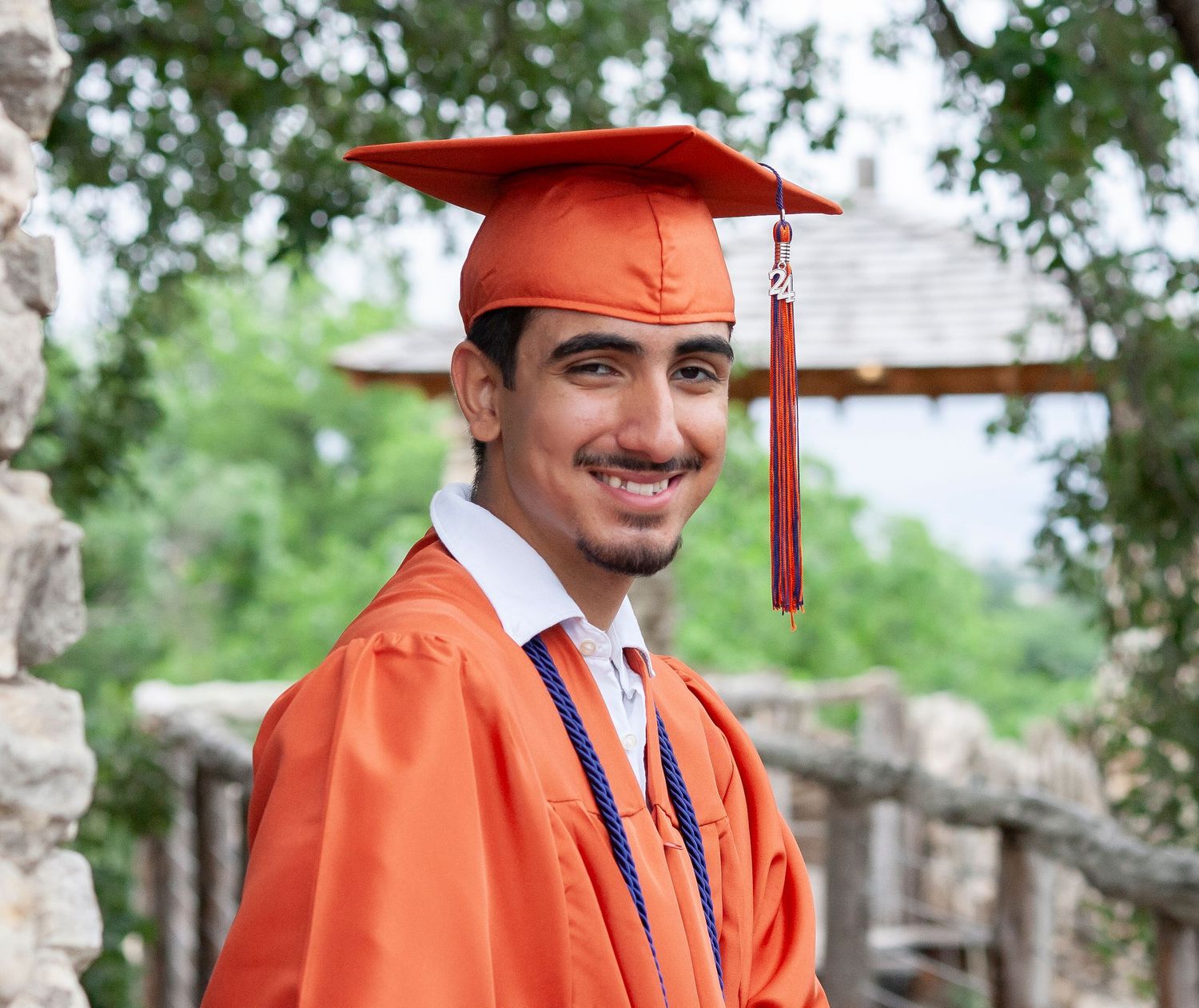 A graduate in orange cap and gown poses near stone architecture.