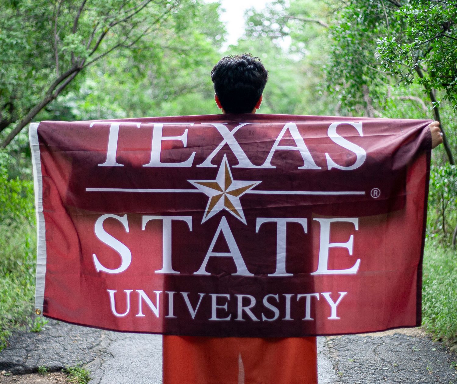A maroon Texas State University banner with white text and star logo held up against a green outdoor background.